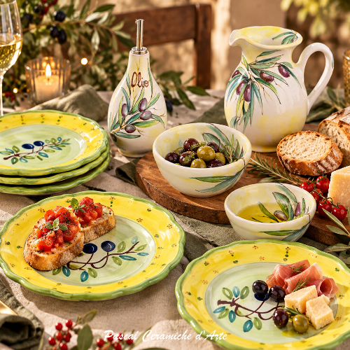 Festive Mediterranean table setting with hand-painted Italian ceramic appetizer plates in yellow and green, olive-themed serving pieces, dipping bowls, olive oil bottle, bruschetta, olives, cheese, and olive oil.